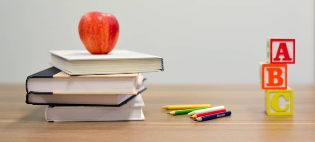 Roter Apfel auf einem Bücherstapel, daneben Buntstifte und gestapelte Holzwürfel mit den Buchstaben A, B, C auf einem Holztisch