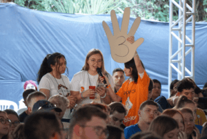 Junge Frau hält Mikrofon, daneben hebt jemand ein Pappschild mit Hand und der Zahl 3 hoch; umgeben von sitzendem Publikum