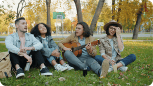 Vier junge Menschen sitzen im Herbstpark auf dem Rasen, einer spielt Gitarre, alle singen oder hören zu.