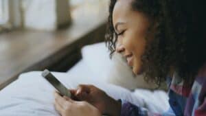 Lächelnde junge Frau mit lockigem Haar liegt auf einem Bett und schaut auf ihr Smartphone