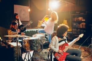 Vier junge Frauen musizieren in einem Proberaum: Schlagzeug, Akustikgitarre, Saxofon und rote E-Gitarre