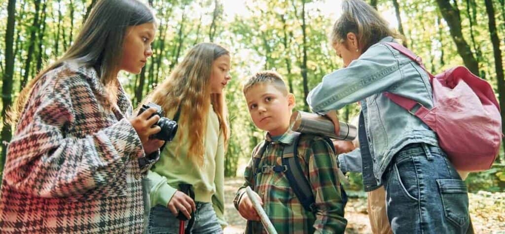 Vier Kinder im Wald, eines hält eine Karte, eines eine Kamera, eines eine Thermoskanne; Sonne scheint durch die Bäume.